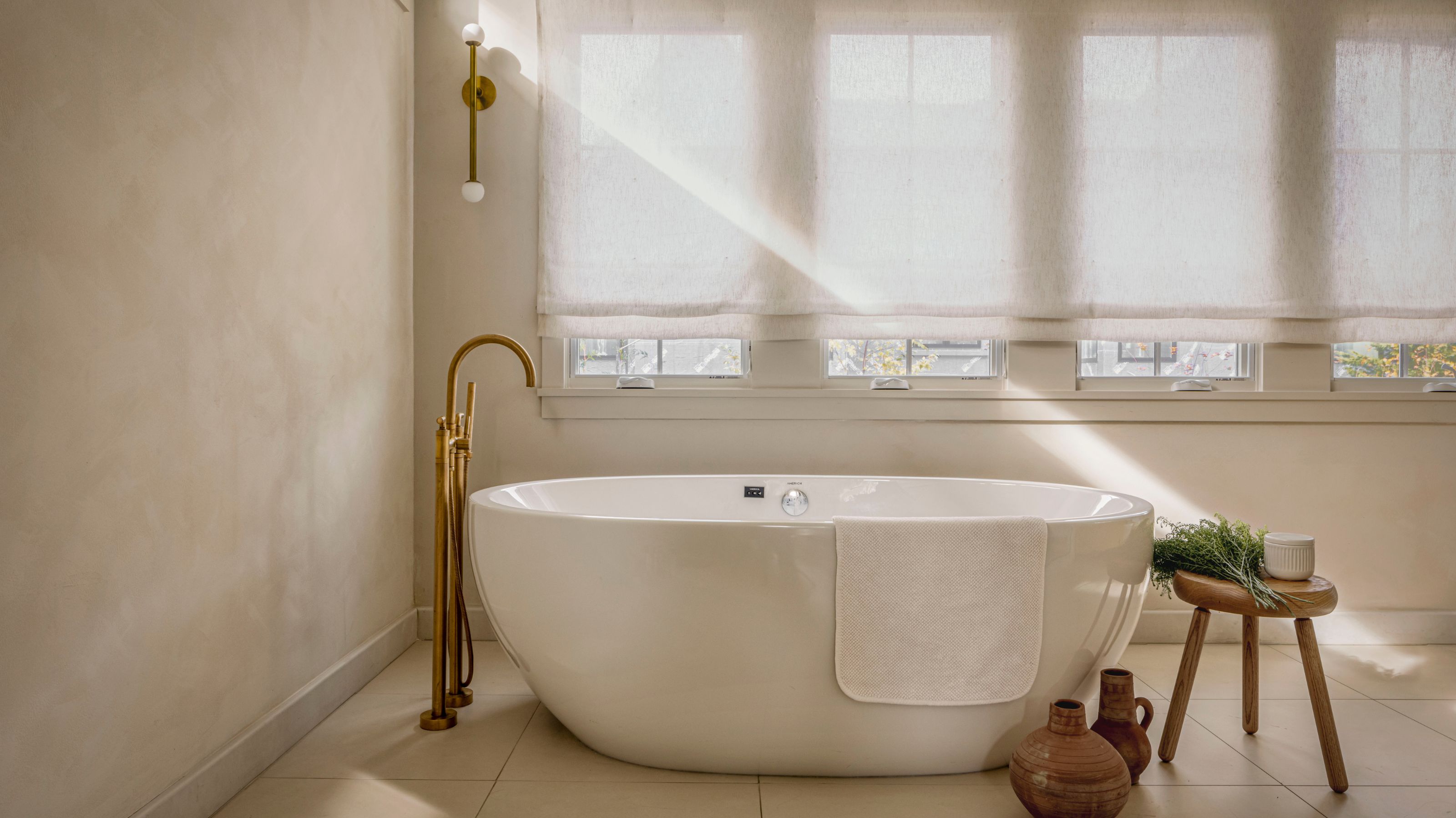 A master bathroom with a large tub and a stool with plants placed next to it. You can also see the tap leaning into the bathtub as well as a golden wall sconce in the background.