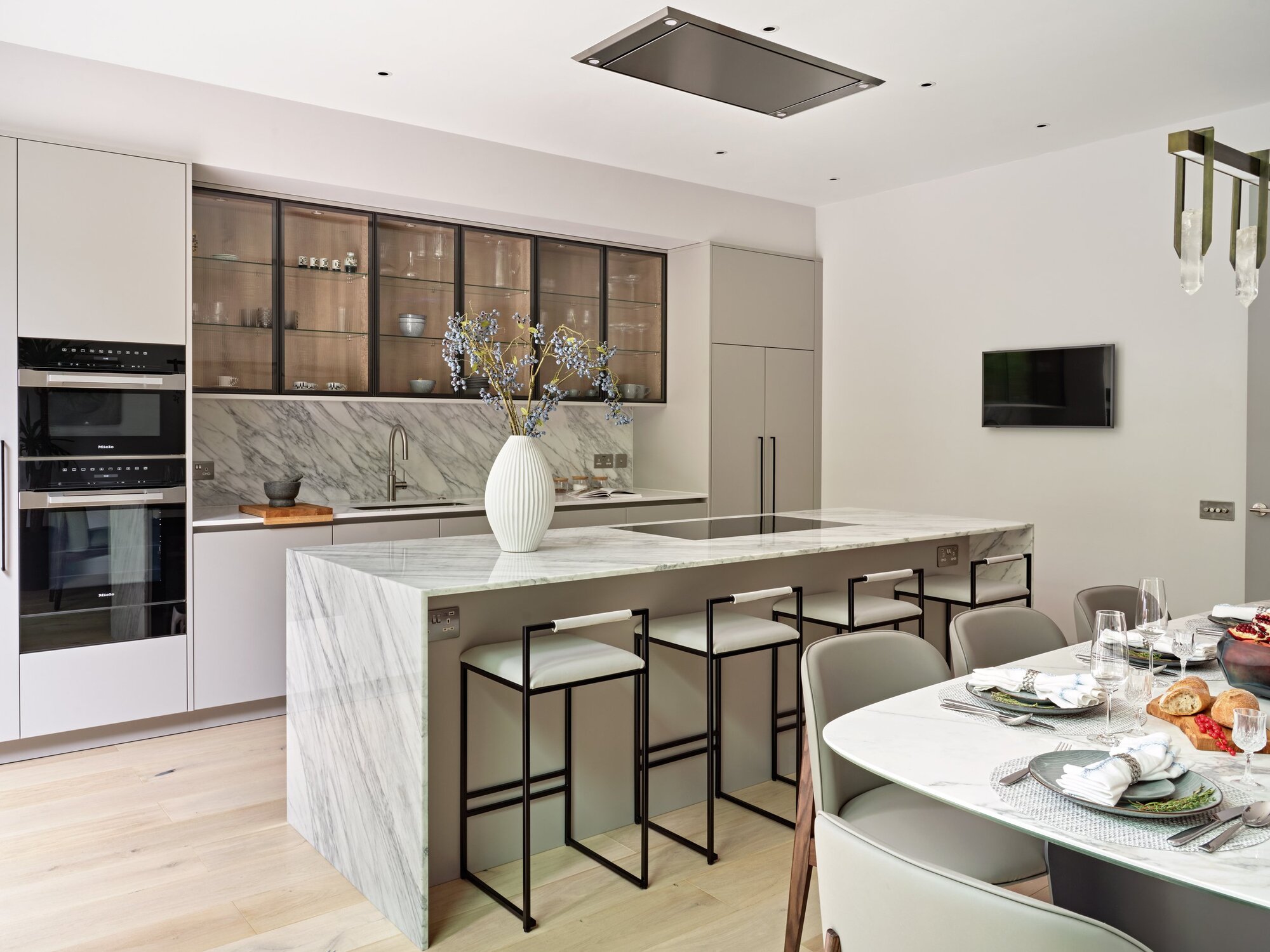 modern kitchen with white cabinets, marble kitchen island, upper cabinets with glass fronts, white and black bar stools, marble dining table, timber floors with vase on the counter