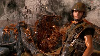 A still from the movie Starship Troopers. On the right we see a soldier dressed in armor and wering a helmet who is covered in alien blood. In the background you can see a giant, bug-like alien monster that has exploded, with orange alien blood everywhere.