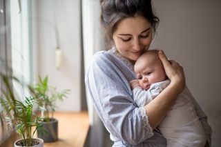 mother holding newborn baby in front of a window at home