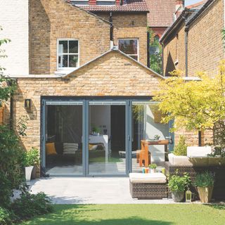 The back garden of a semi-detached house with a bit of grass and a grey patio with rattan furniture on it