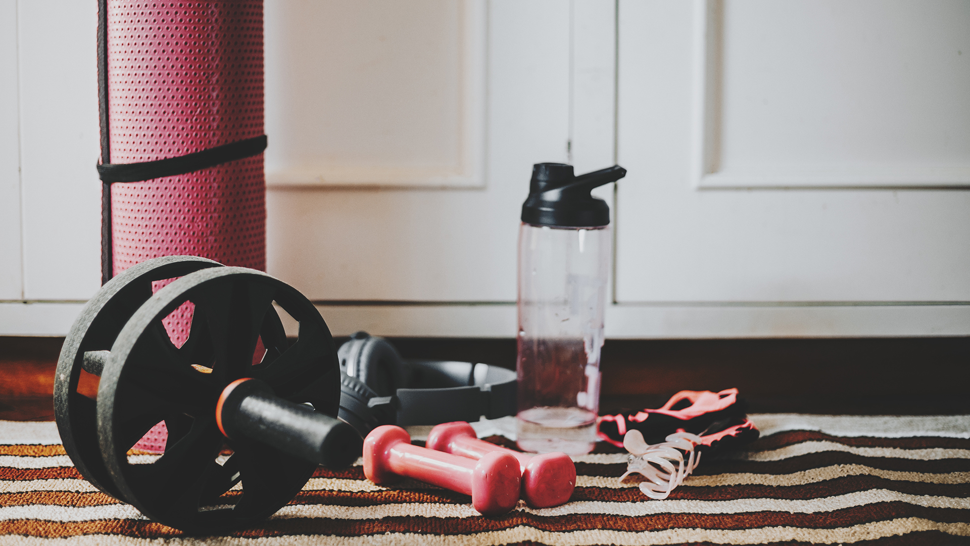 Bedroom floor with exercise equipment including a yoga mat, mobility wheel, weights and water bottle