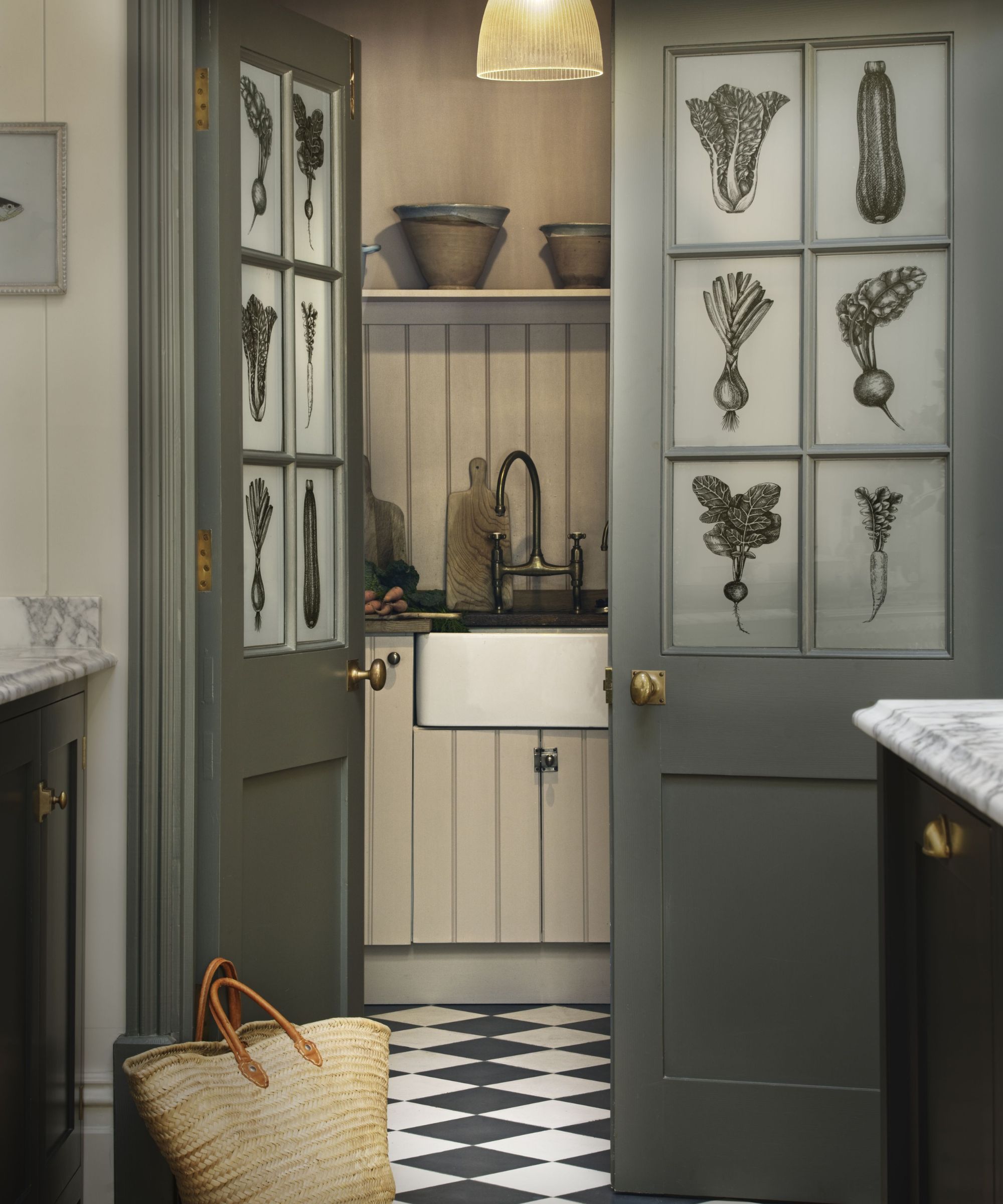 A view into a pantry or utility space through green-painted doors with frosted glass panels decorated with botanical illustrations. The space features a white farmhouse sink, a black tap, wooden shelving with ceramic bowls, and vertical panelled walls.