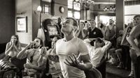 Black and white image of a crowded pub with people reacting emotionally, some seated and others standing near a slot machine