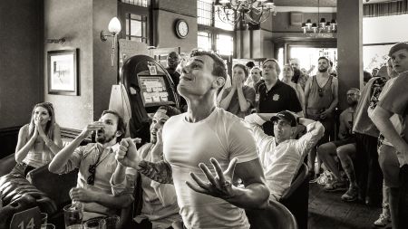 Black and white image of a crowded pub with people reacting emotionally, some seated and others standing near a slot machine