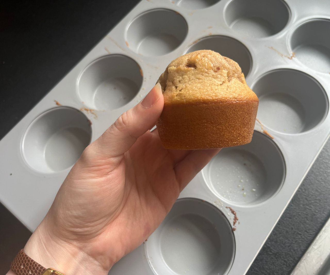 A muffing with a golden base being held above a grey 12-hole muffin tin