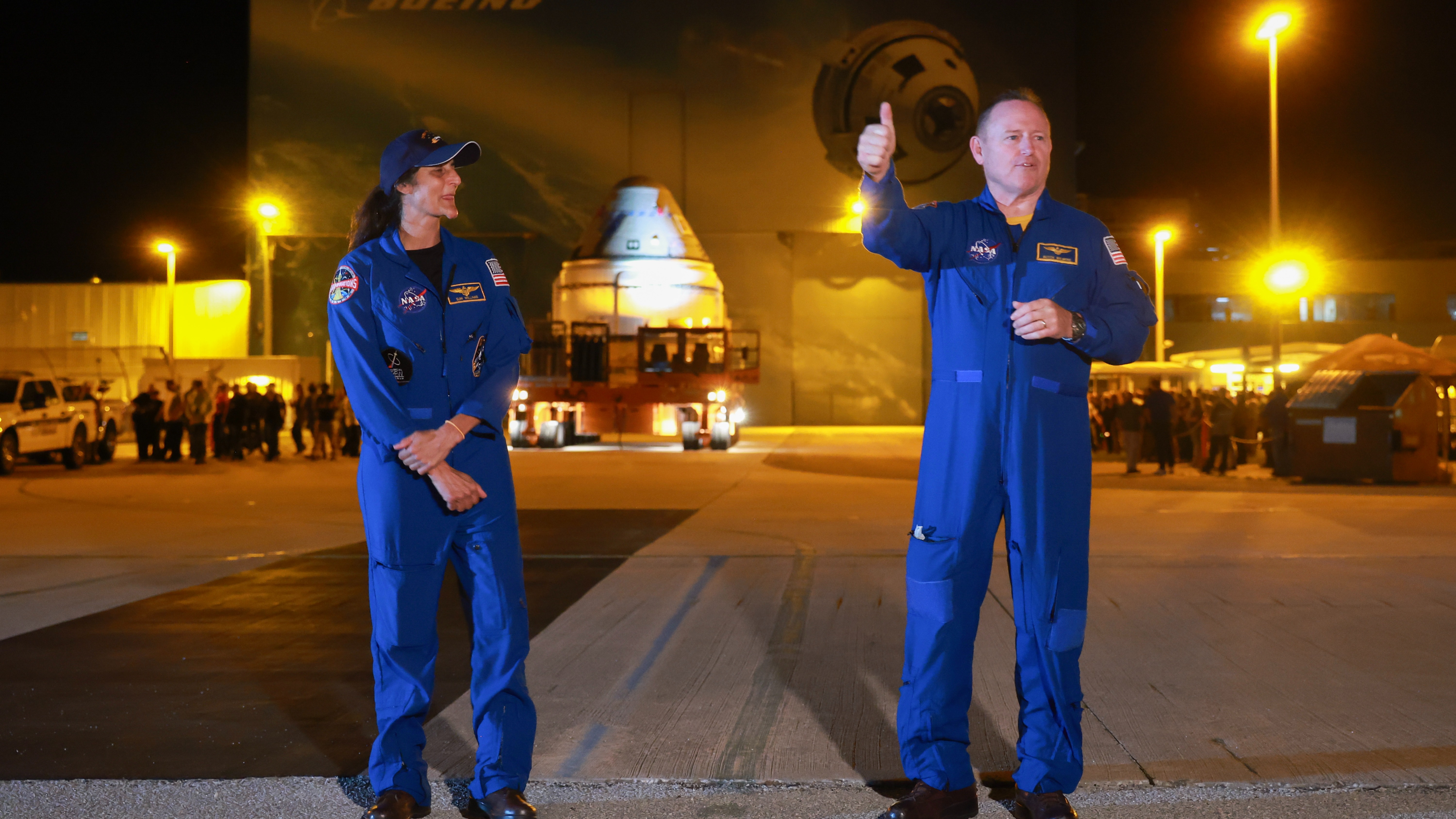 two astronauts in flight suits in the dark. the one on the right flashes a thumbs-up. far in the background is a cone-shaped spacecraft on a trailer, in front of buildings