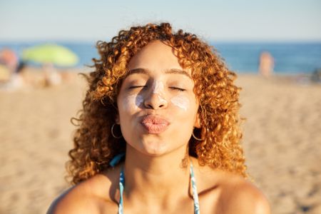 A woman with brown curly hair is sitting on a beach, she has suncream on her cheeks. She is facing the camera with her eyes closed and her lips puckered. 