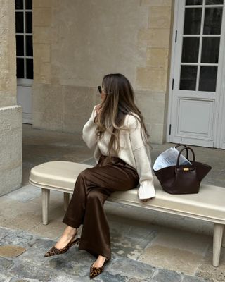 @oliviafaeh vestindo um suéter creme, calça de seda marrom, sapatos slingback com estampa de leopardo e uma bolsa Demellier New York marrom