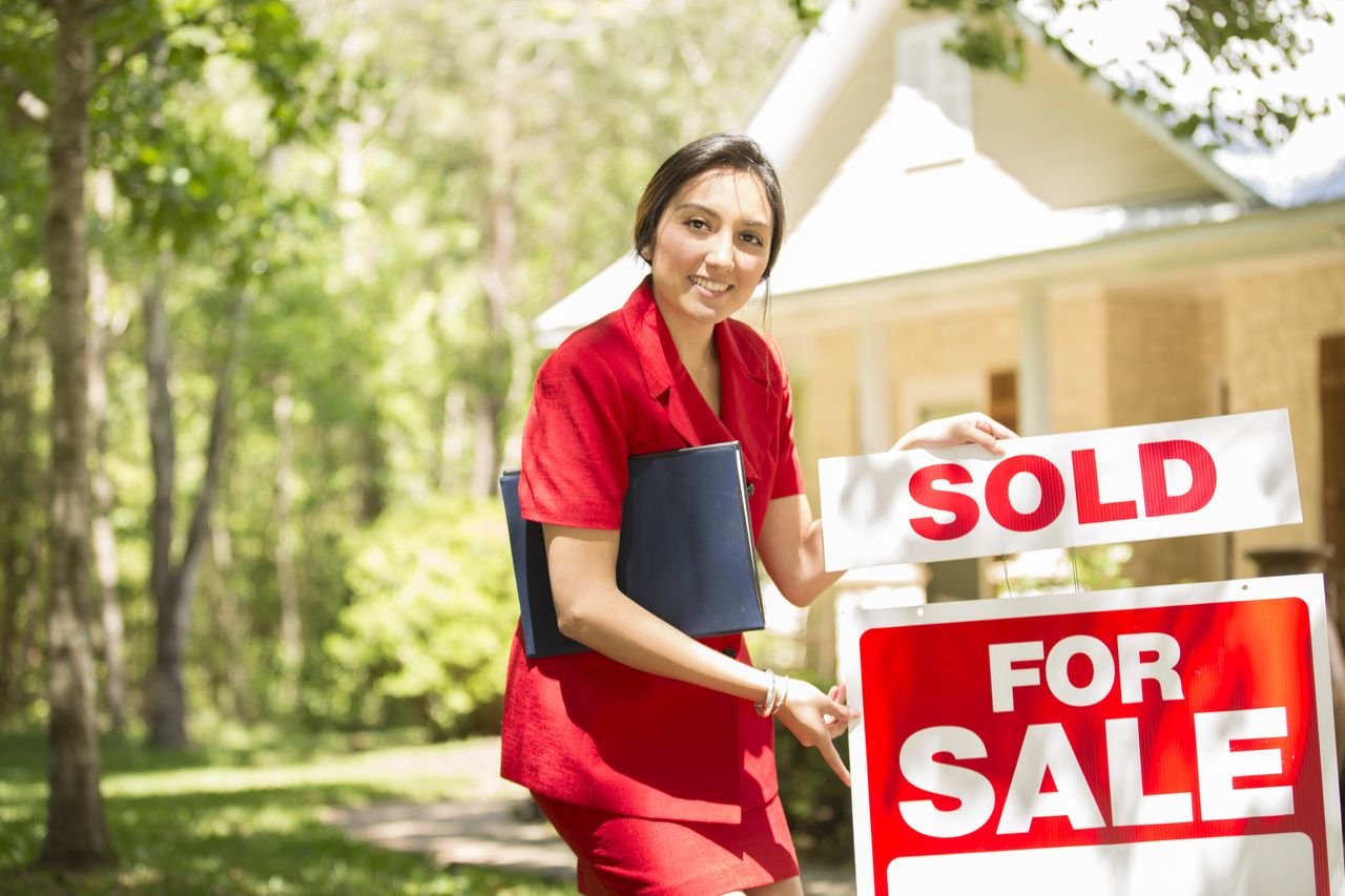 Real Estate Agent with home for sale. She places a sold sign in yard. Home in background in spring or summer season