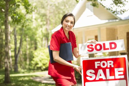 Real Estate Agent with home for sale. She places a sold sign in yard. Home in background in spring or summer season