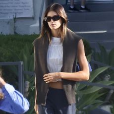 Kaia Gerber walks down the street wearing navy trousers and burgundy ballet flats with a white short sleeve blouse and a brown knit over her shoulders.