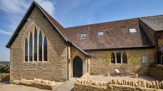 A converted Cotswold stone chapel with tall Gothic arched windows, a dark arched door, tiled roof with skylights and a gravel courtyard with dry stone walls.