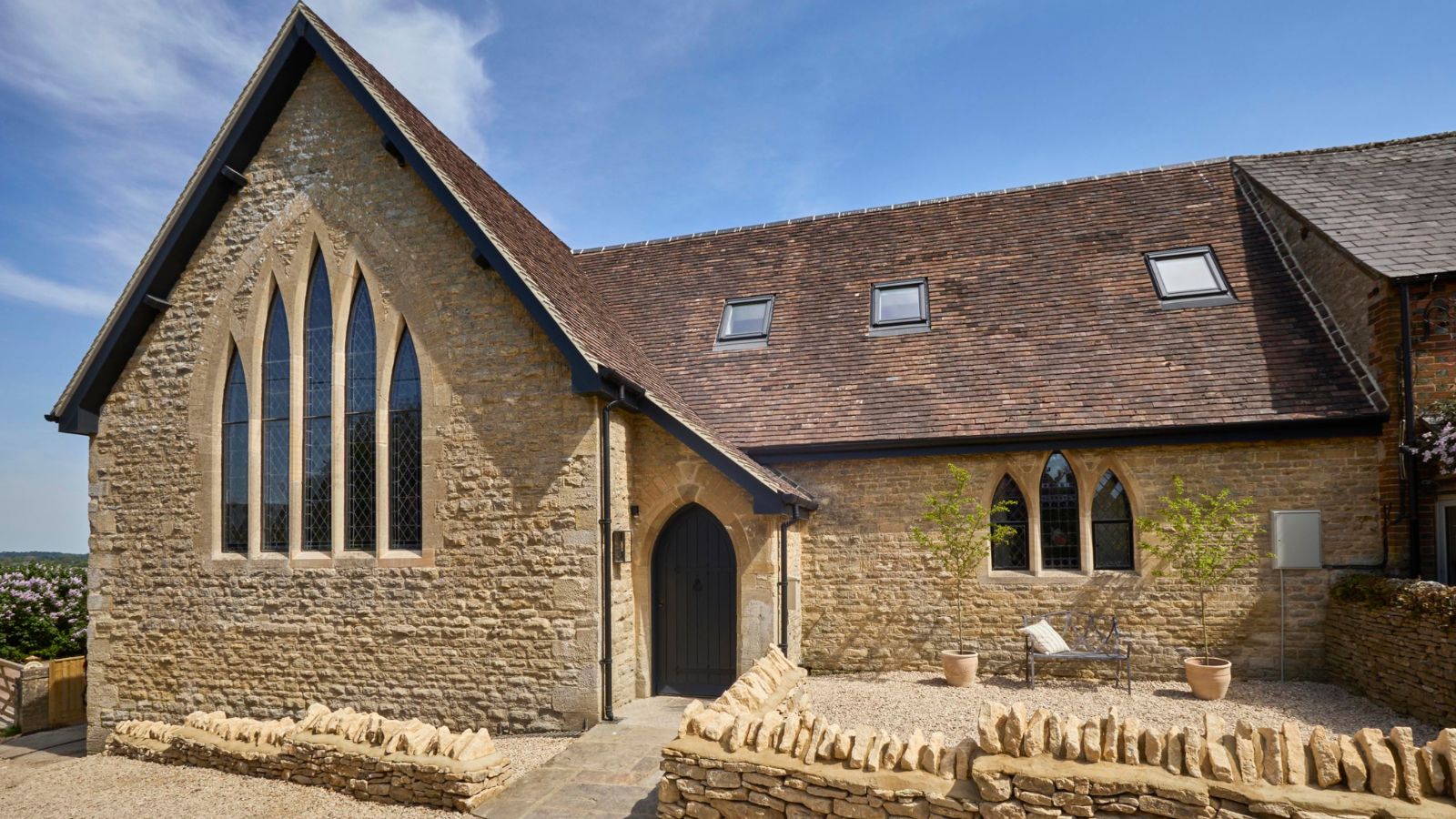 A converted Cotswold stone chapel with tall Gothic arched windows, a dark arched door, tiled roof with skylights and a gravel courtyard with dry stone walls.