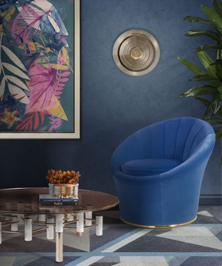 Bold MCM blue lounge with blue velvet curve chair, round metallic coffee table, dark blue walls, and multi-colored leaf print on wall.