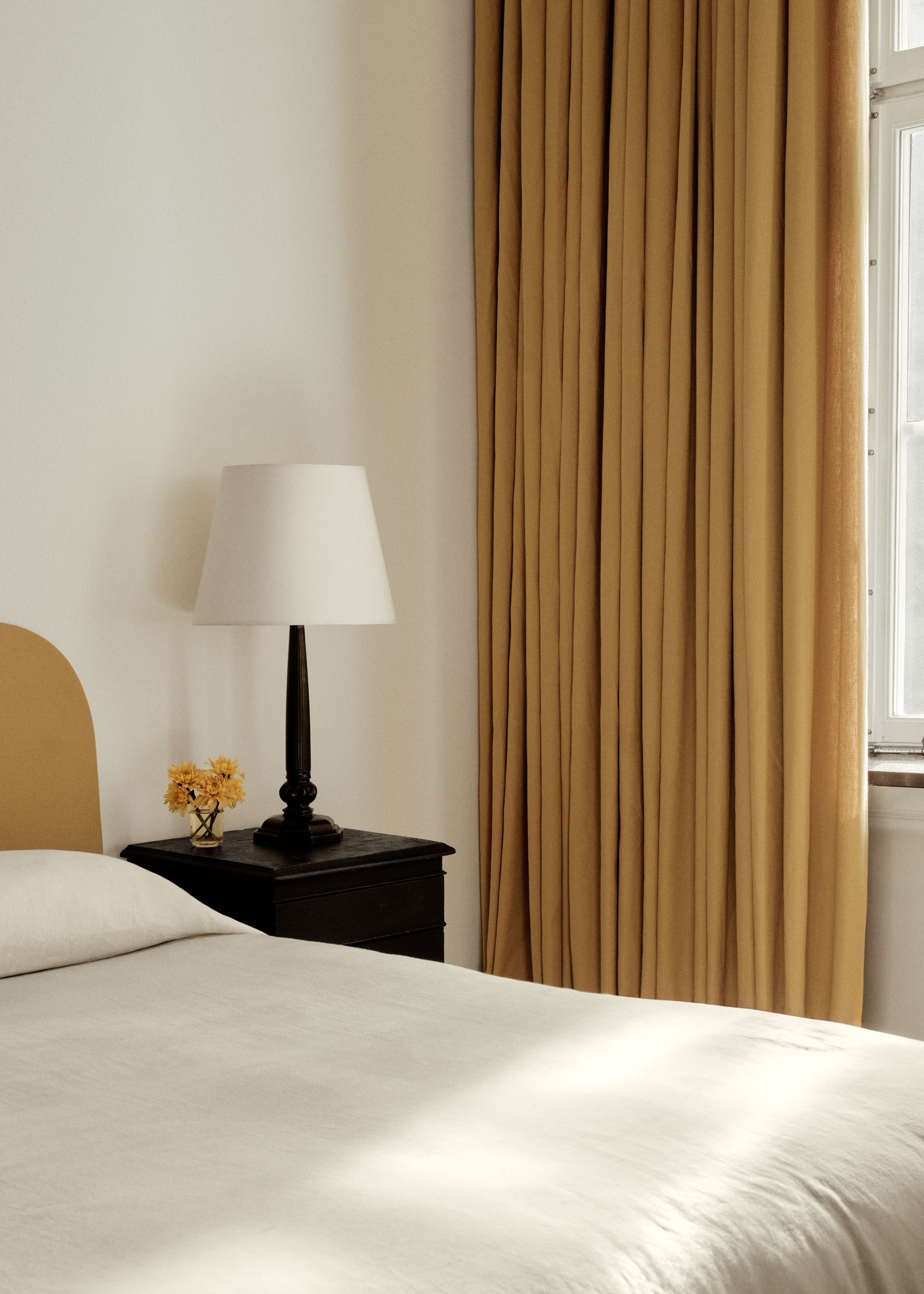 Corner of a neutral bedroom with mustard yellow curtains, white walls, soft white bedding, and a black nightstand with a lamp.
