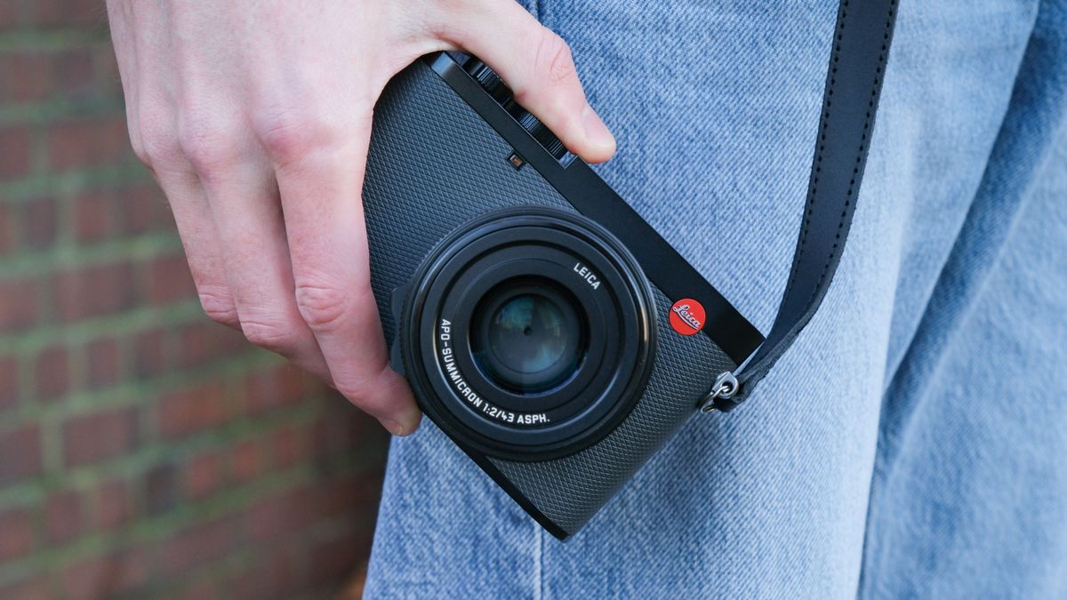 A Leica Q3 43 camera with a strap attached held in a hand by a person's side