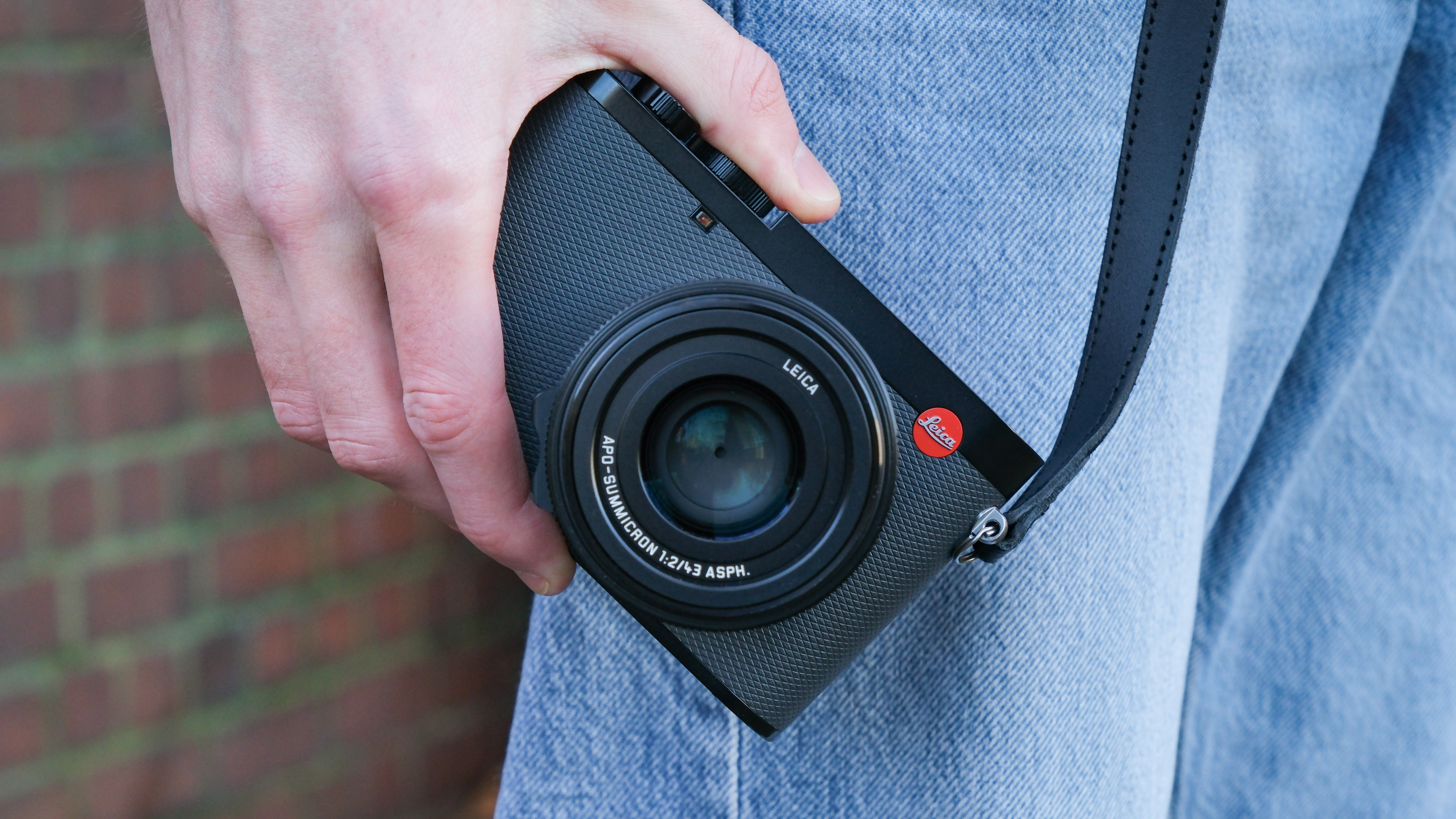 A Leica Q3 43 camera with a strap attached held in a hand by a person's side