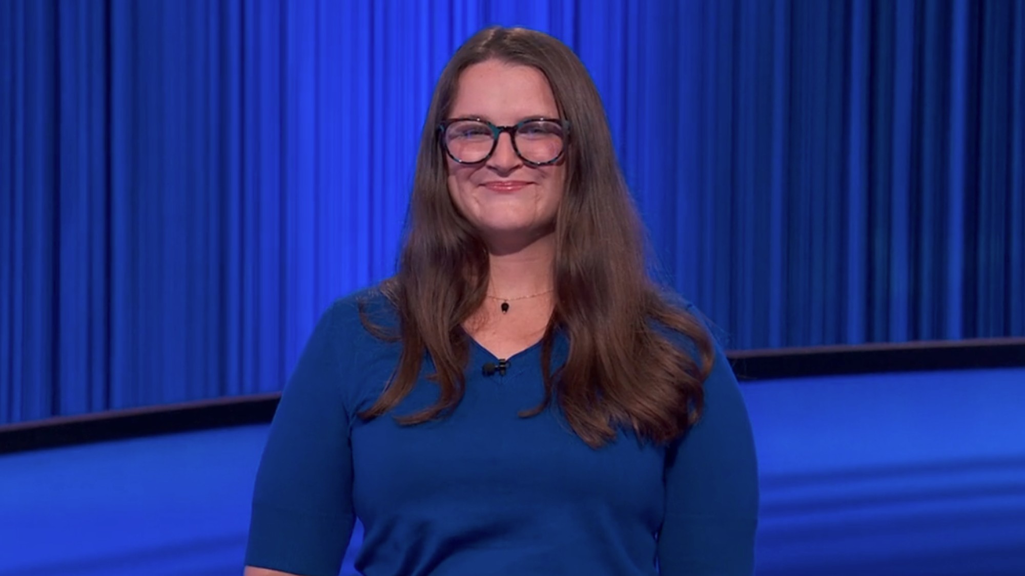 Megan Whelan getting introduced on Jeopardy, wearing black glasses and a blue sweater.