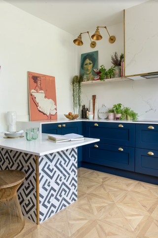 A blue kitchen with blue cabinetry, white and blue tiled kitchen island, brass kitchen wall lighting, light wood-effect flooring and framed wall art decor