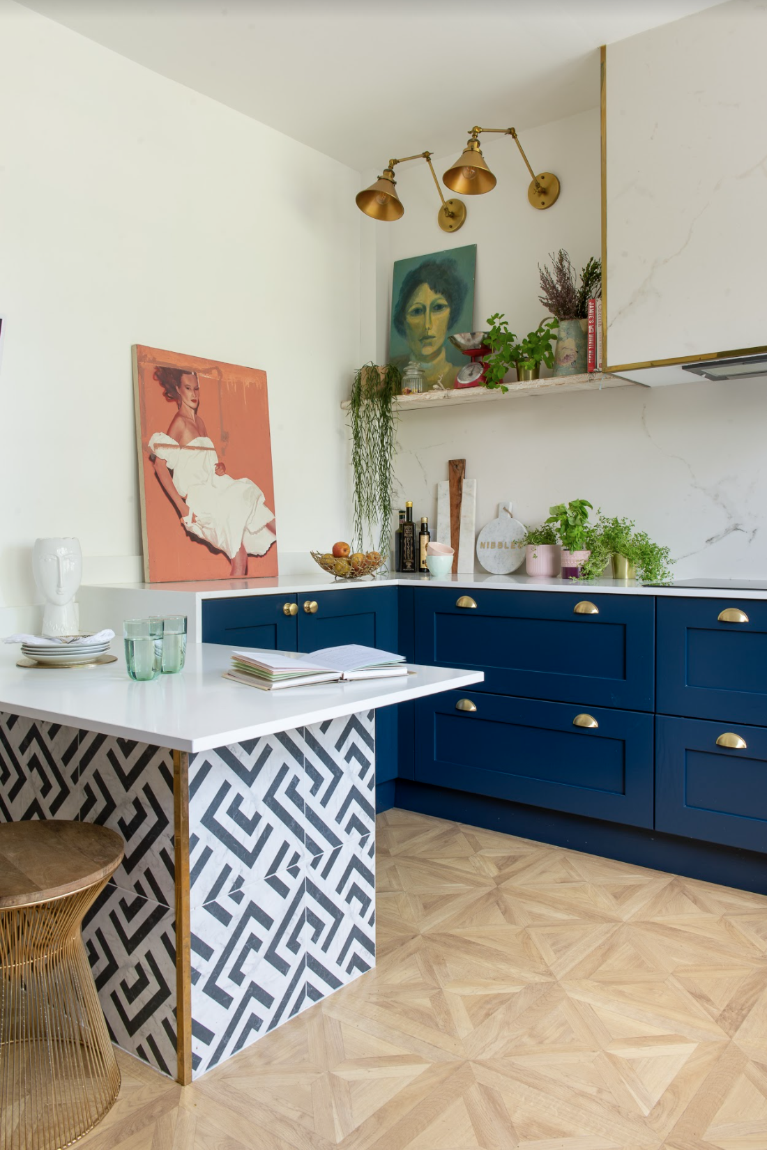 A blue kitchen with blue cabinetry, white and blue tiled kitchen island, brass kitchen wall lighting, light wood-effect flooring and framed wall art decor