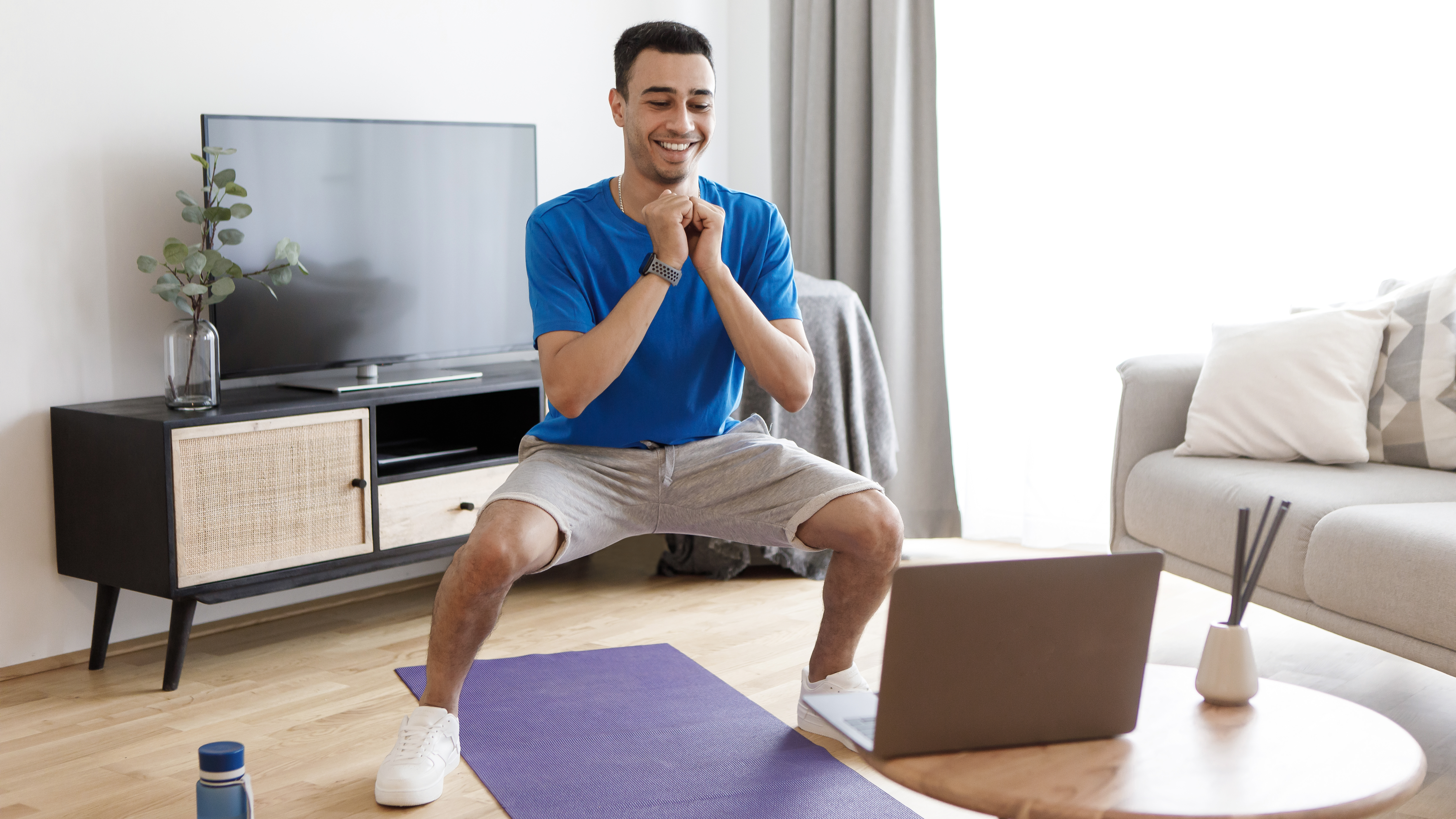 A man performs a squat at home while looking at a laptop on a coffee table. He is smiling and we see a couch and TV behind him. 