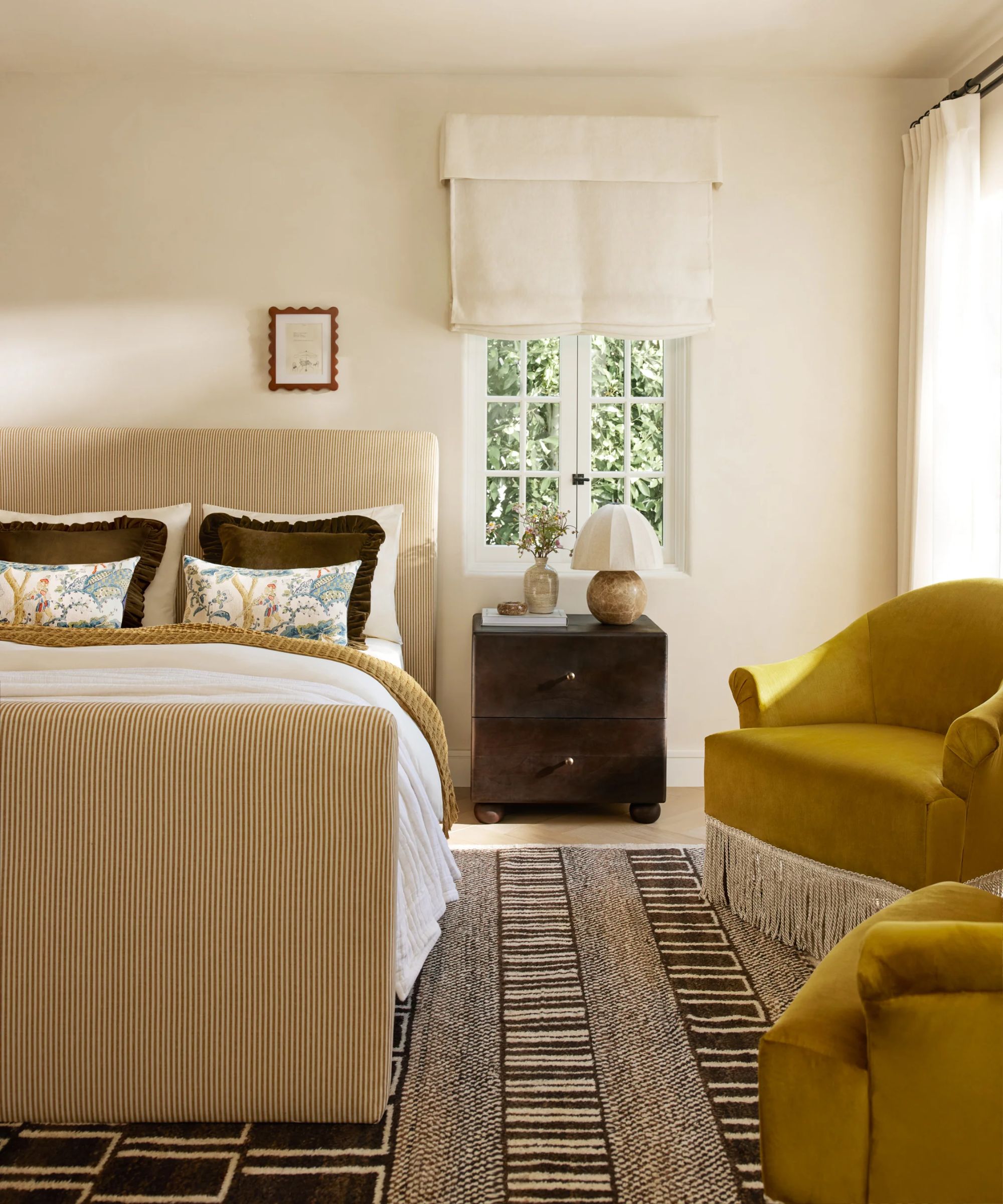 neutral bedroom with a striped ochre upholstered bed, dark brown nightstand, mustard armchairs with fringed trim