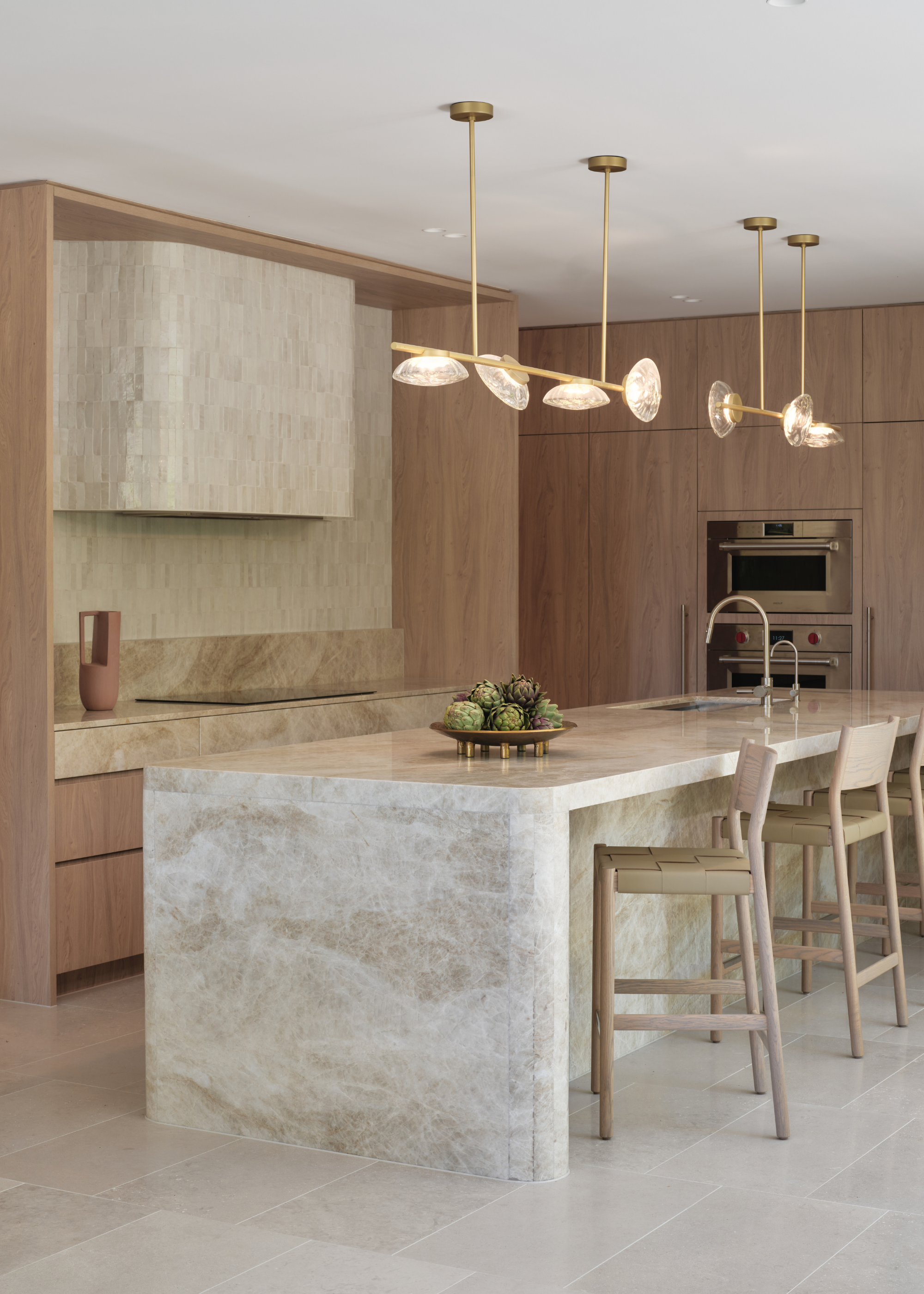 A bright kitchen with a stone island, cream chairs, a structural overhead light, wood cabinets, a tiled range hood and backsplash, and a bowl of artichokes