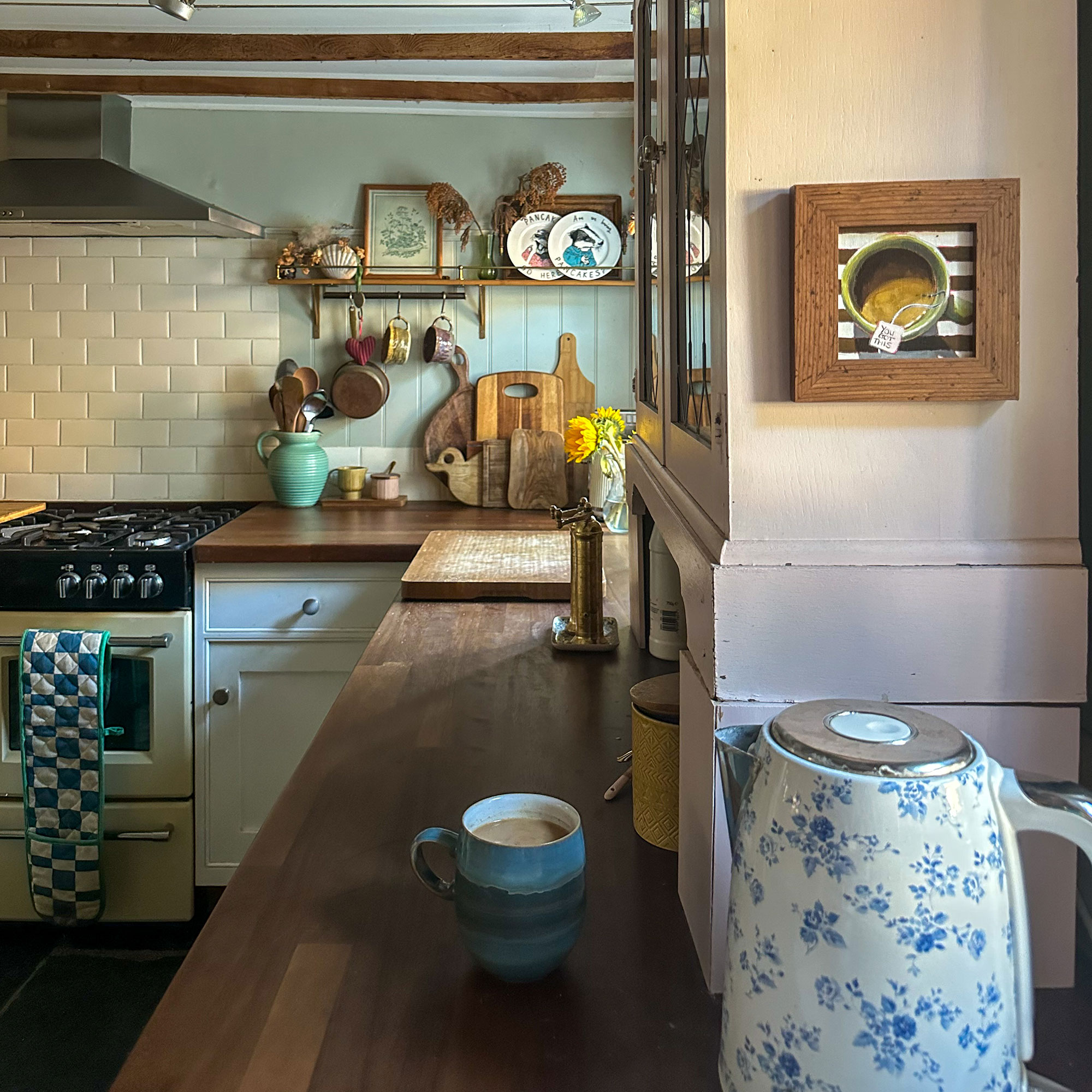 Country kitchen with wooden worktops, ceiling beams and small framed artwork above blue and white floral patterned kettle