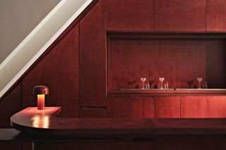 A red-drenched, minimalist bar inspired by Bauhaus features a sculptural, mushroom-shaped red table lamp on a curvilinear counter, behind of which we see three transparent wine glasses next to drink bottles.