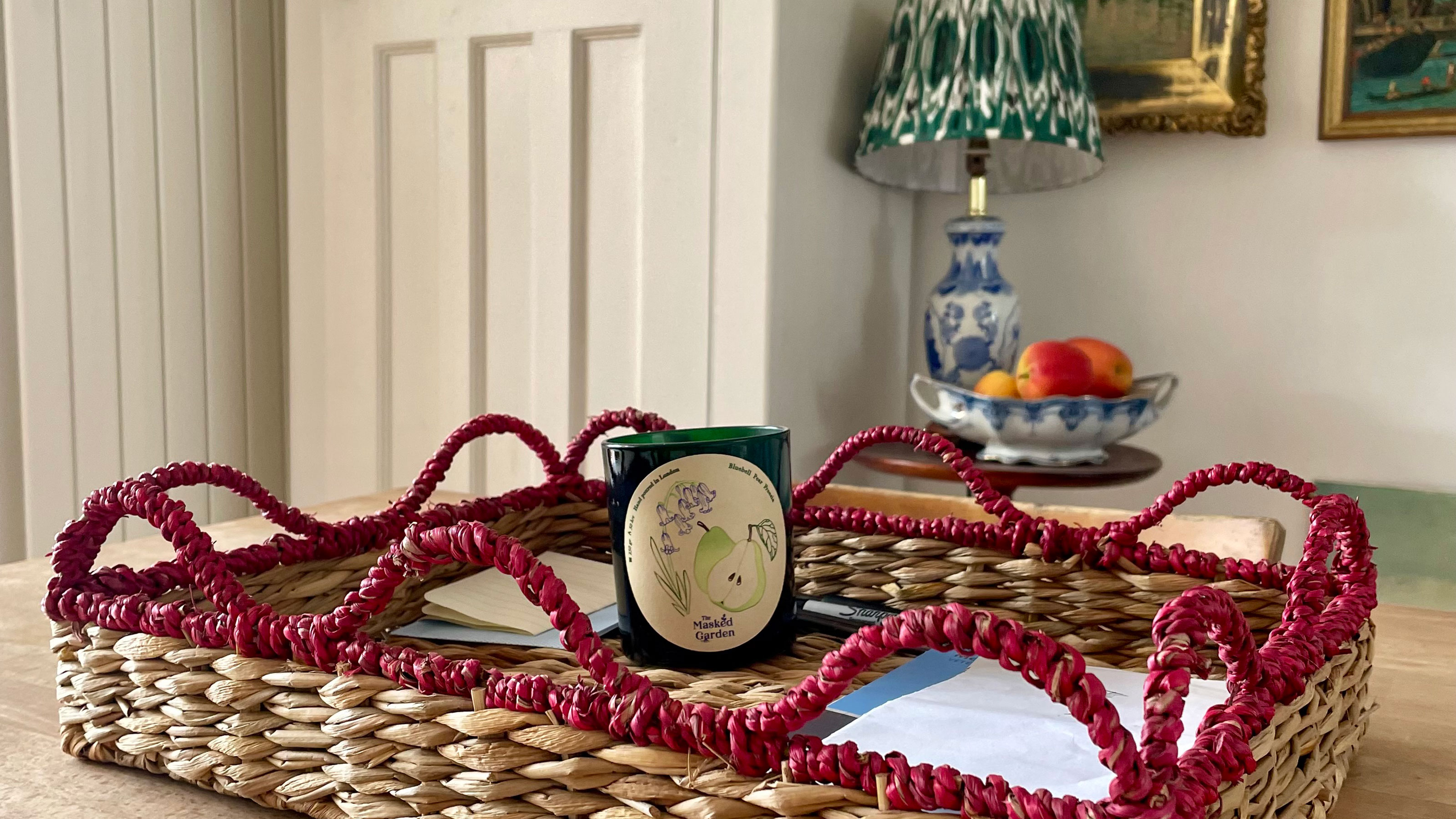 Woven seagrass tray with red wavy trim holding a candle in front of lamp and bowl of fruit