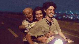 Fabietto rides a motorbike smiling as a man and woman sit behind holding onto his waist smiling.