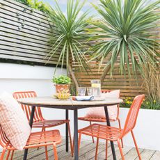 a courtyard garden with horizontal slated fencing and palm trees alongside coral coloured metal dining chairs and a wooden table