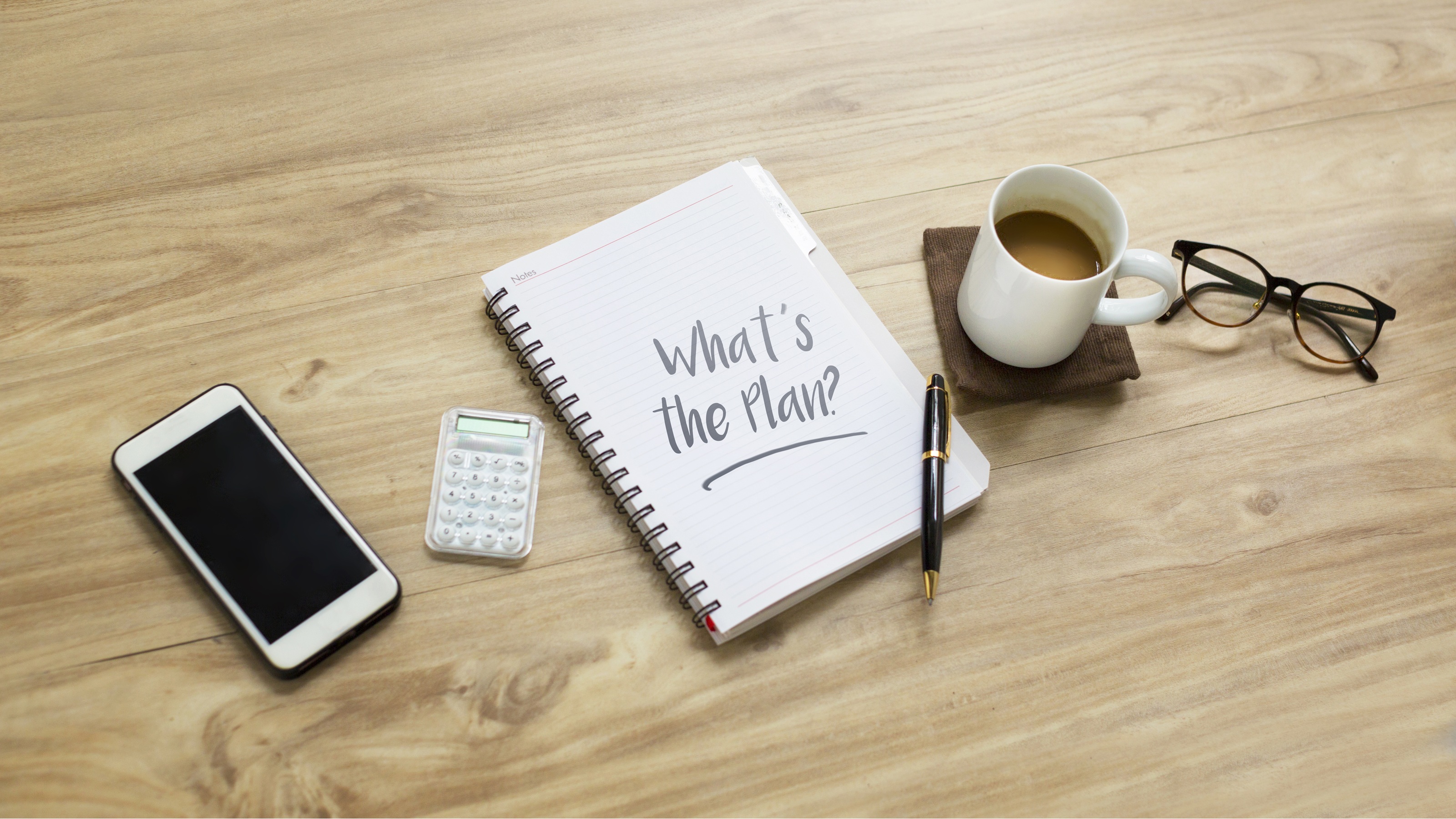A phone, calculator, cup of coffee, eyeglasses and a notebook with "What's the plan?" written on a page are arranged on a table.