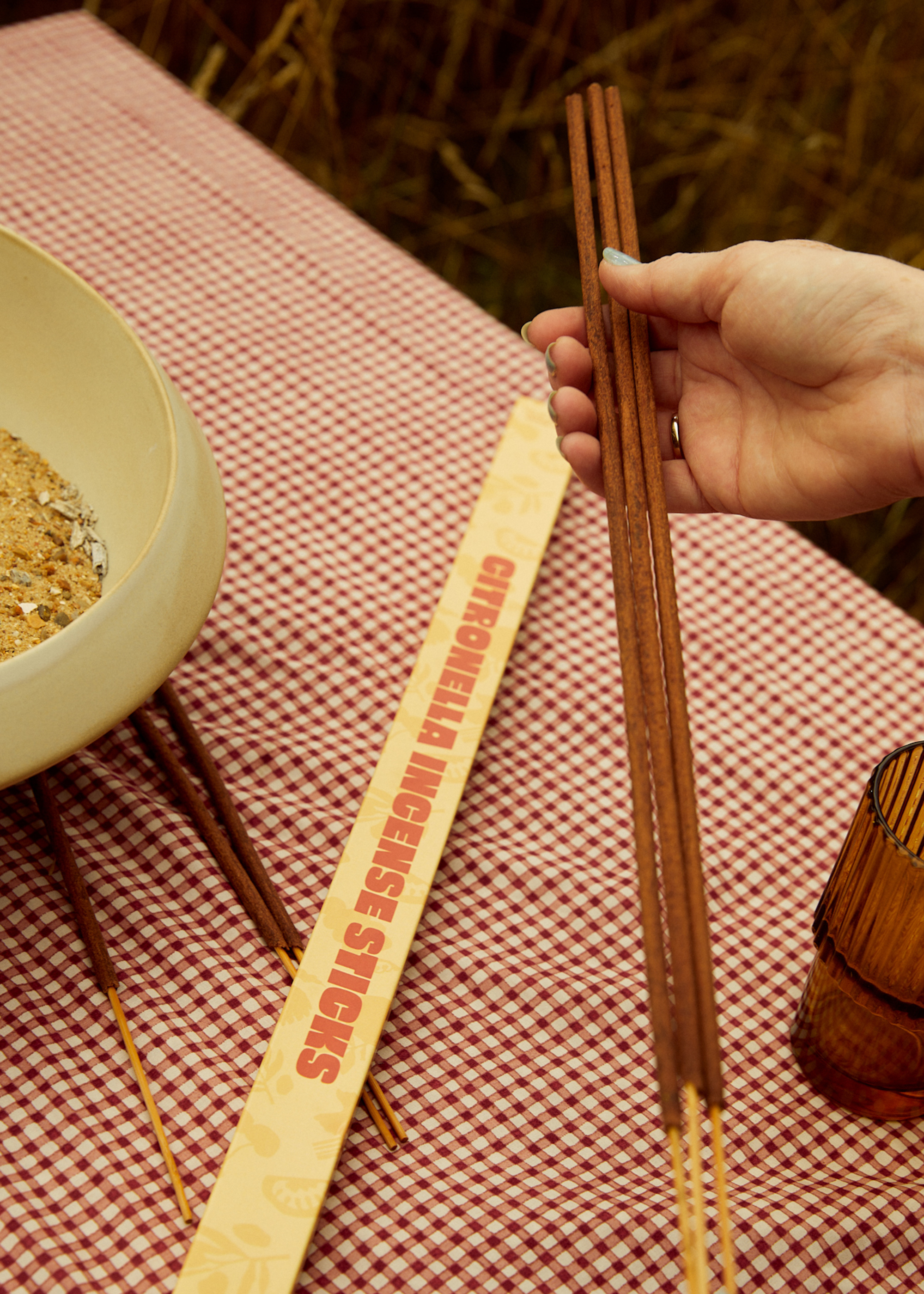 A hand holding three long incense sticks over a table dressed in gingham tablecloth with an amber glass and an incense bowl partially in frame