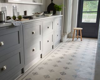 Kitchen with vinyl flooring with a tiled pattern, grey cupboards, white marble countertop, white tiles, light wood stool