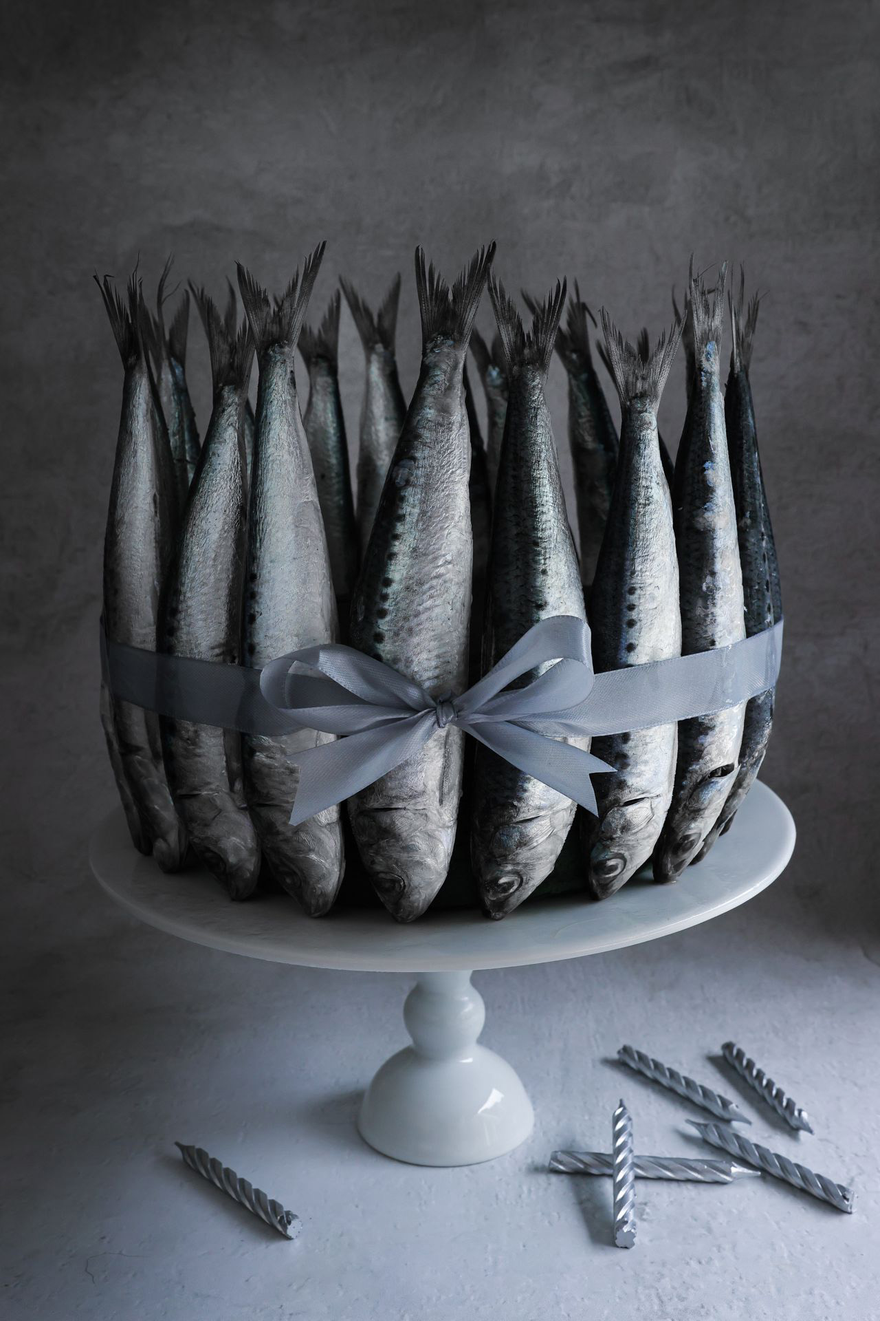 A cake stand displays vertically arranged fish tied with a silver ribbon, resembling a cake. Gray candles lie nearby