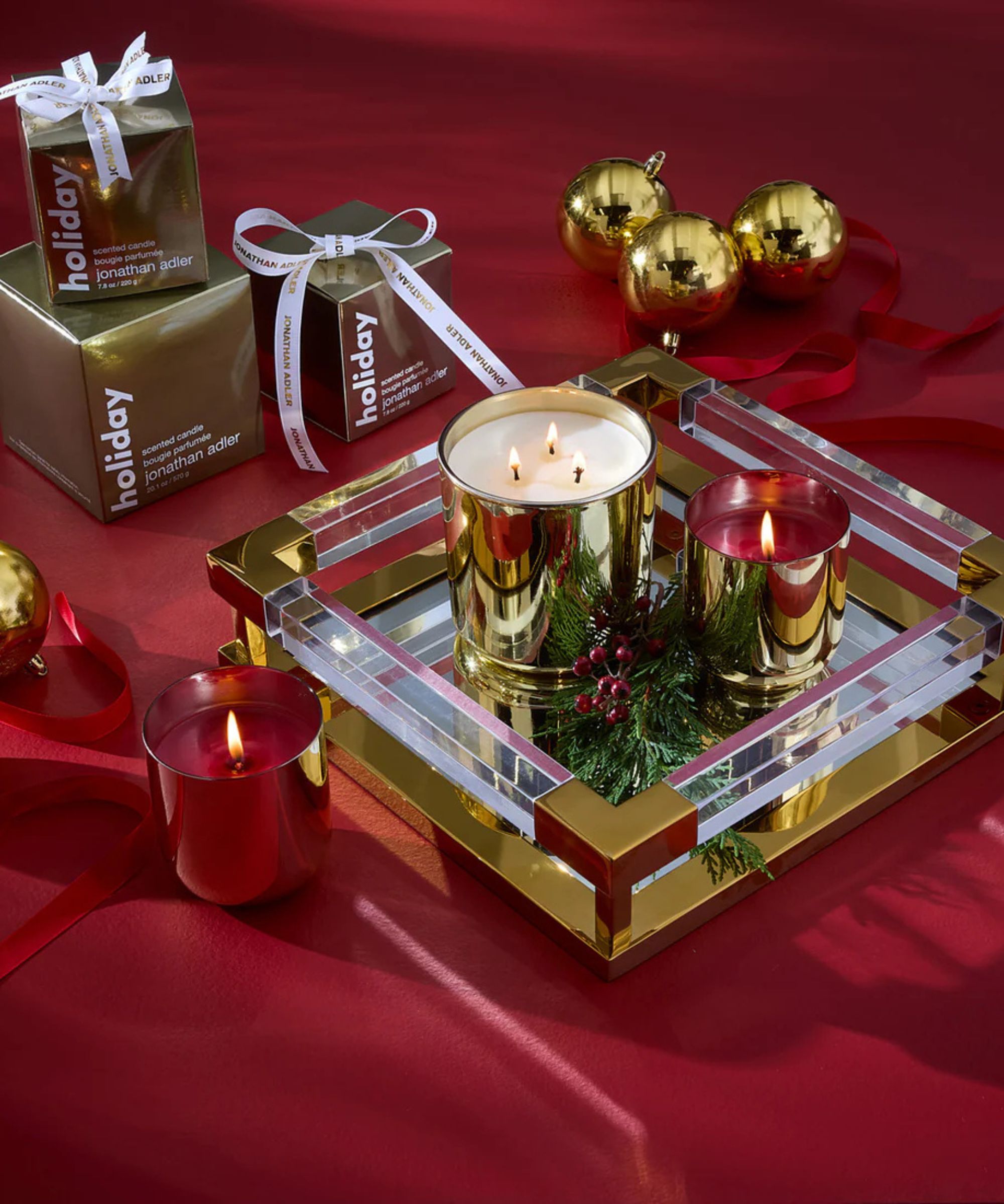 A luxury holiday still life featuring gold and red metallic Jonathan Adler candles, evergreen sprigs, and gold gift boxes on a red backdrop