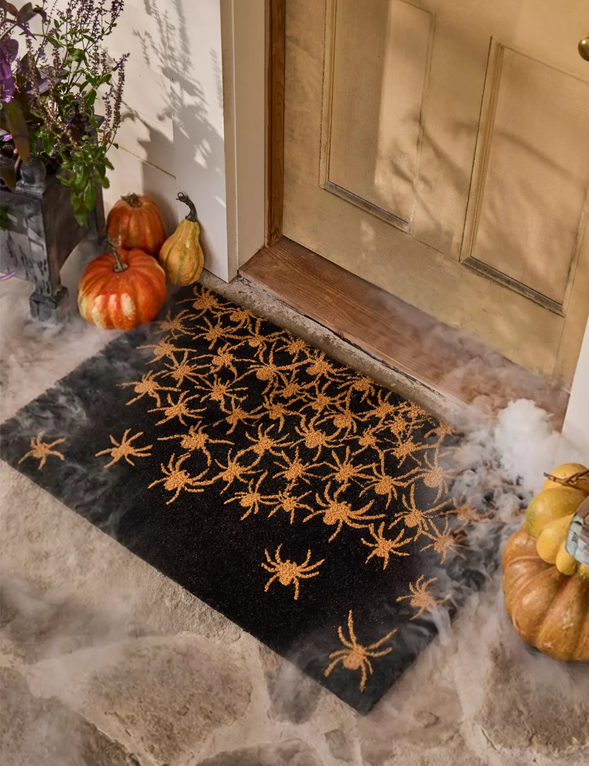 Spider-motif black-and-orange doormat flanked by stacks of pumpkins on a stone porch.