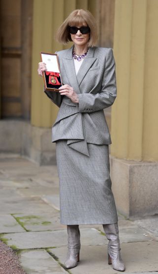 Anna Wintour wearing a gray suit and amethyst necklace with black sunglasses and holding a box with a red medal inside and standing outside on a stone sidewalk