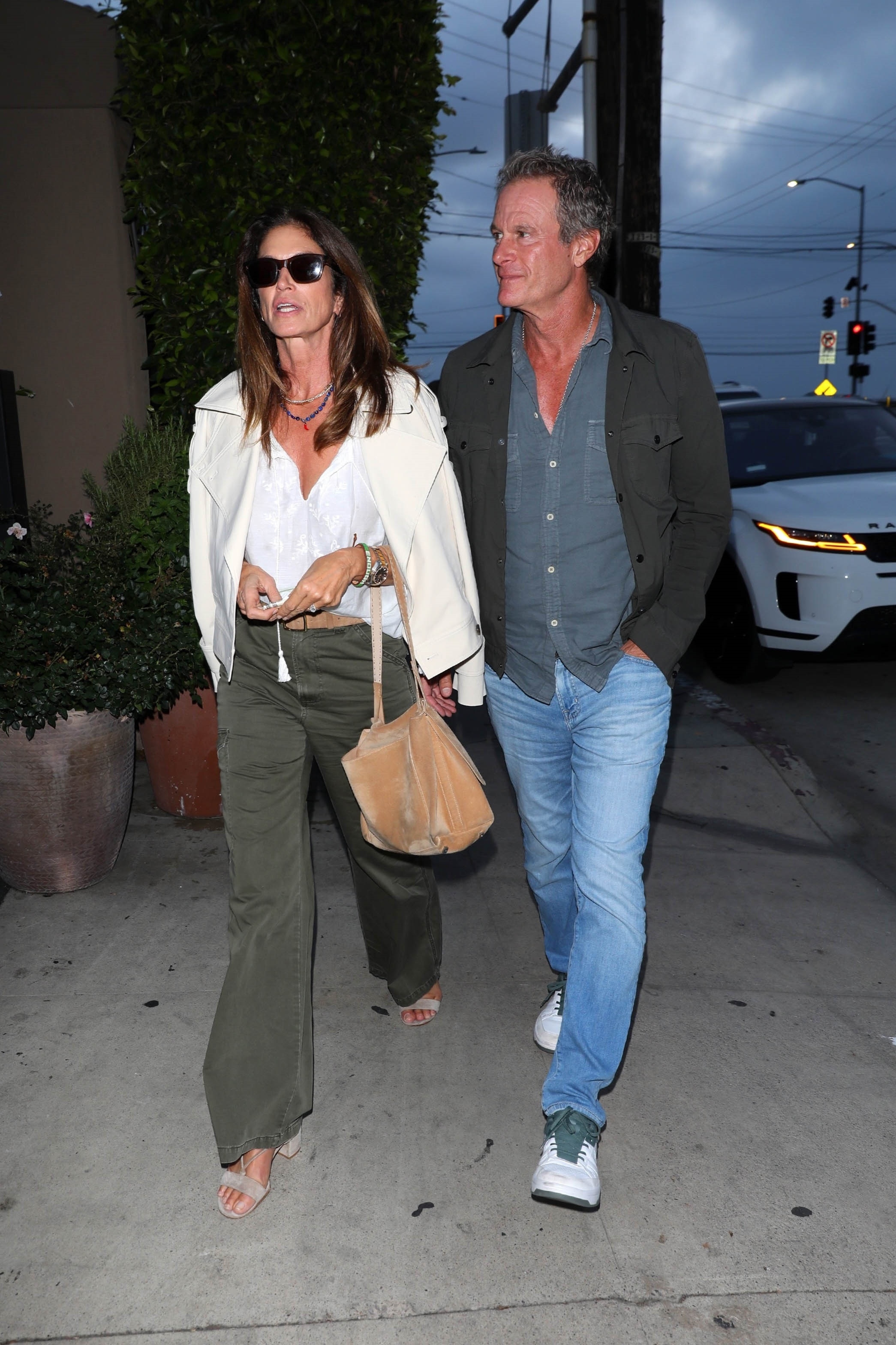 Cindy Crawford walks down the street wearing khaki jeans with beige suede sandals and a white blouse.