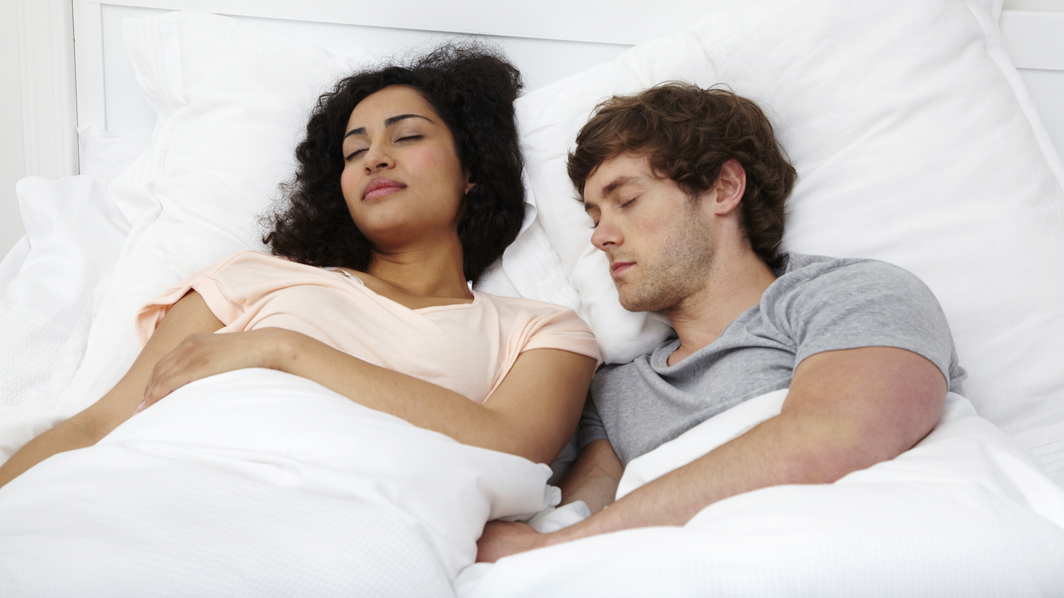 Images shows a couple asleep together in bed, resting on white pillows