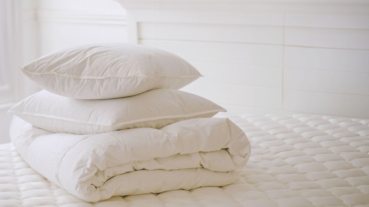 Duvet and two pillows piled on top of a bare mattress in a white bedroom