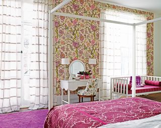 Girls' bedroom decor with bold floral wallpaper, a white dressing table and crimson patterned throw on a white four poster bed.