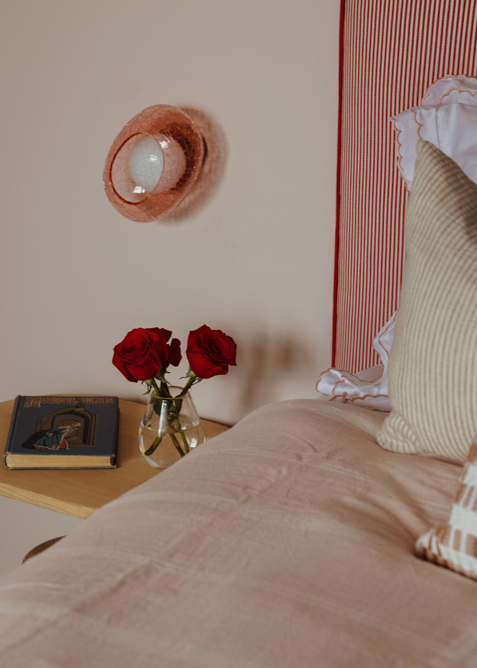 A blushy bedroom with soft pink accents in the form of bedding and a glass wall sconce, alongside a striped headboard, a wooden side table with a vase of roses and a hardcopy of Arabian Nights