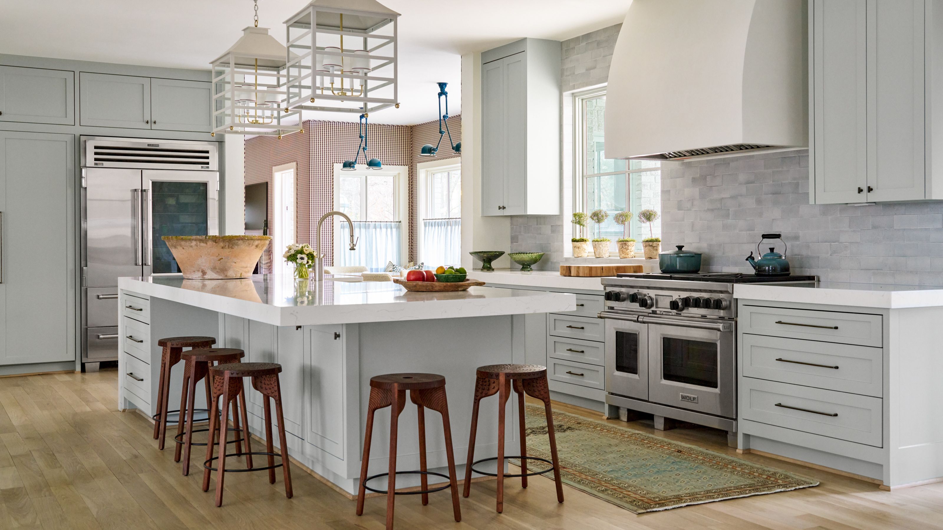A spacious, light-filled modern kitchen featuring pale blue-gray shaker cabinets and white countertops. A large kitchen island dominates the center, providing seating with four wooden stools. The kitchen includes a professional stainless steel range, a large white hood, and a subtle gray subway tile backsplash, with two white lantern pendant lights hanging overhead.