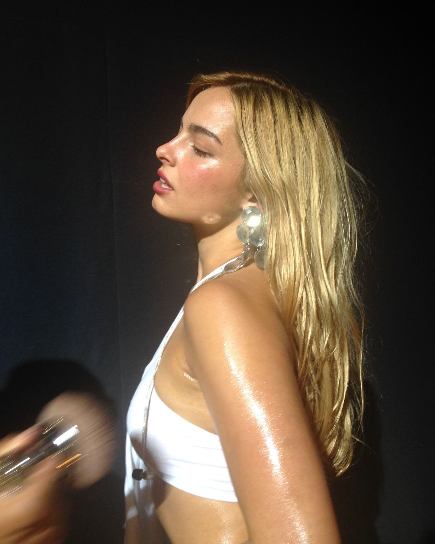 Addison Rae with blonde hair and glowing skin, wearing a white crop top and having a makeup artist apply a body shimmer to her arms