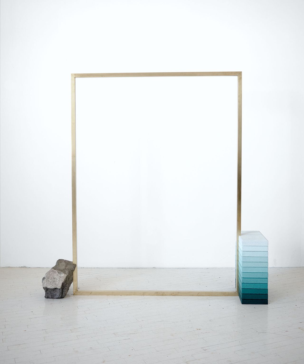 A conceptual sculpture features a golden frame, a grey stone, positioned at its left-end bottom corner, and a pile of gradient seafoam green bricks.