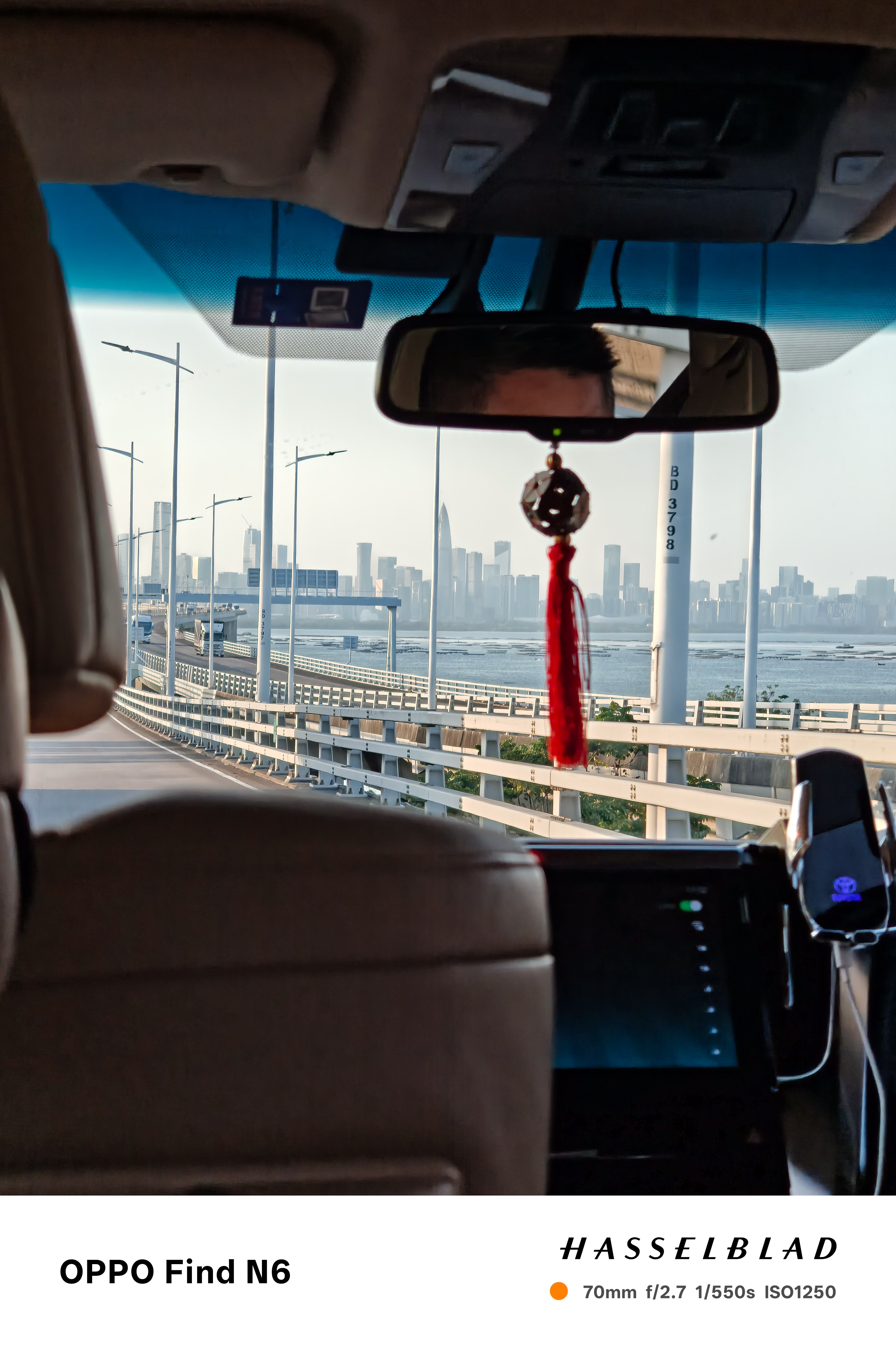 A view from the interior of a car driving across a long bridge toward a modern city skyline. The foreground shows the car's tan leather headrest and a red tassel hanging from the rearview mirror. Outside, the bridge&rsquo;s white railings lead the eye toward a hazy cluster of skyscrapers across the water under a clear, bright sky.