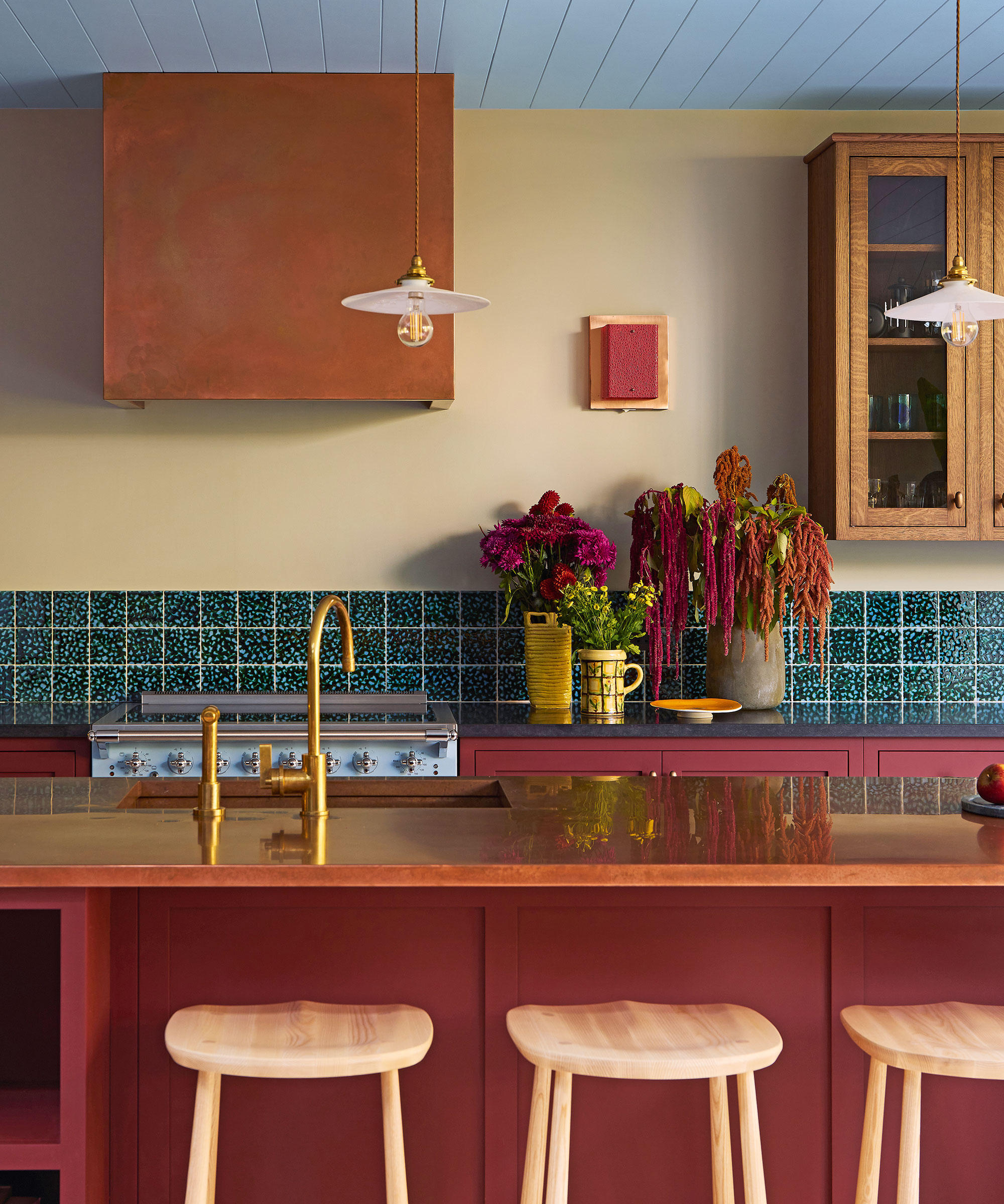 A contemporary kitchen designed by Tom Morris featuring a bold red island with light wood stools and a striking copper range hood against a pale yellow wall. The space is accented by a vibrant teal tiled backsplash, a light blue paneled ceiling, and elegant gold fixtures, with lush floral arrangements providing a touch of organic texture.